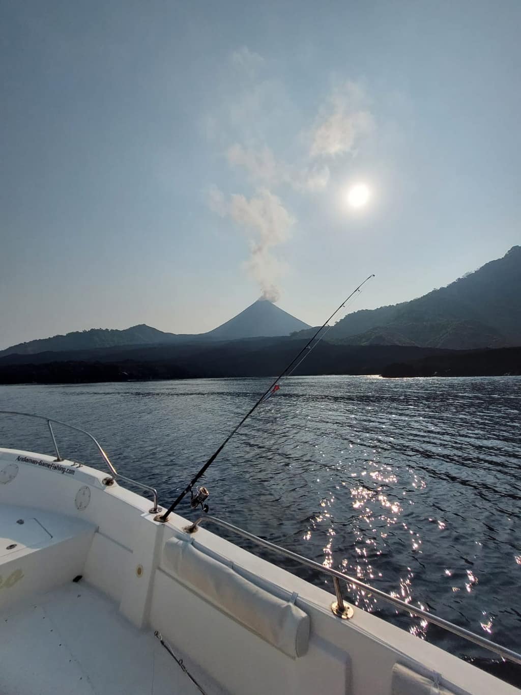 The Barren Island volcano venting a plume of ash and steam, seen from a boat with the sun behind it